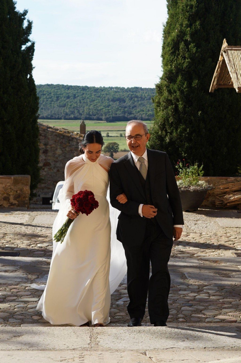 Bride walking arm in arm with her father across a stone courtyard